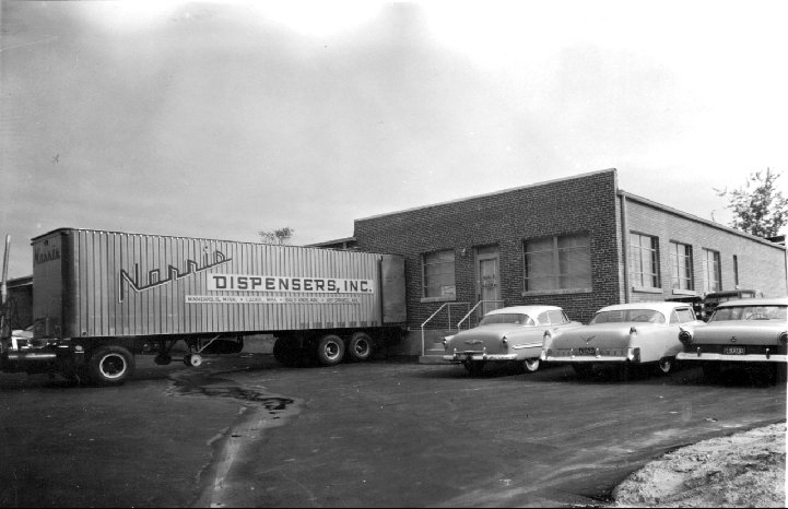 Black-and-white photo of a Norris Dispensers, Inc. trailer and 1950s cars outside a brick building