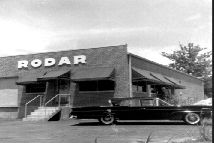 Black-and-white photo of a brick building with a RODAR sign and a vintage car parked in front